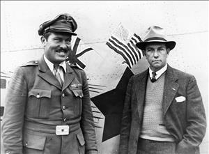 Two pilots stand in front of airplane, one in uniform and one in a suit, both wearing hats